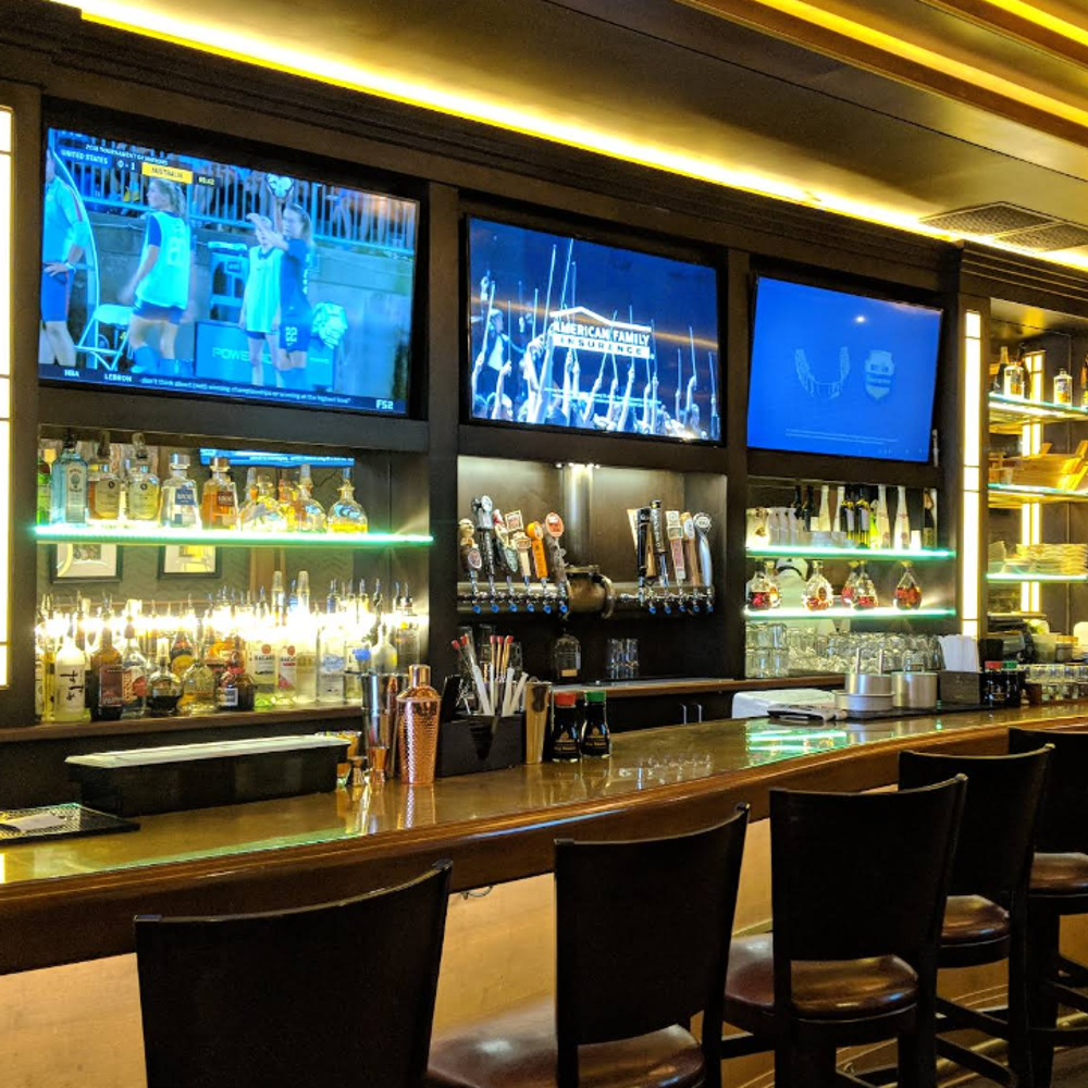 The-1126-Restaurant-Sushi-Restaurant-Columbus A dimly lit bar with a polished wooden counter, leather stools, and multiple TVs above shelves of liquor bottles and beer taps.